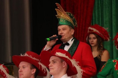 Mann in rotem Jackett und grünem Hut mit goldener Verzierung und Schriftzug 'NCC' spricht in Mikrofon, weitere Personen mit roten Hüten und Federn im Hintergrund.