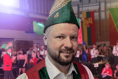 Mann in rotem Jackett und grünem Hut mit goldener Verzierung und Schriftzug 'NCC' spricht in Mikrofon, weitere Personen mit roten Hüten und Federn im Hintergrund.