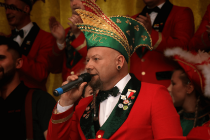 Mann in rotem Jackett und grünem Hut mit goldener Verzierung und Schriftzug 'NCC' spricht in Mikrofon, weitere Personen mit roten Hüten und Federn im Hintergrund.