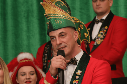 Mann in rotem Jackett und grünem Hut mit goldener Verzierung und Schriftzug 'NCC' spricht in Mikrofon, weitere Personen mit roten Hüten und Federn im Hintergrund.